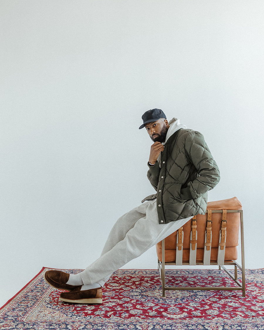 A man in an olive jacket and hoodie sits atop a leather chair.