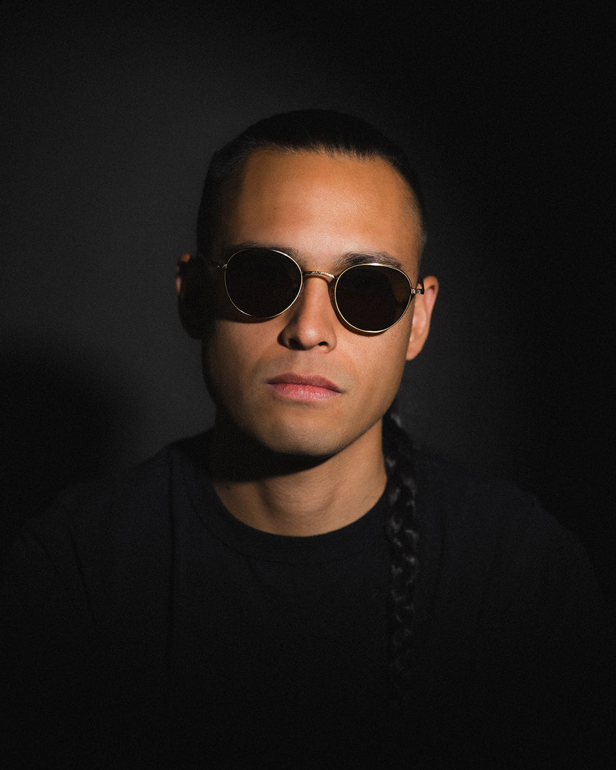 A man in round sunglasses poses in front of a black background.