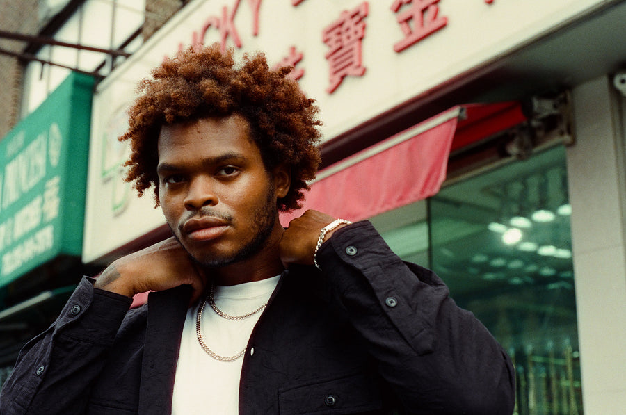 A man with bright chain necklaces and a black shirt on looks into the camera.