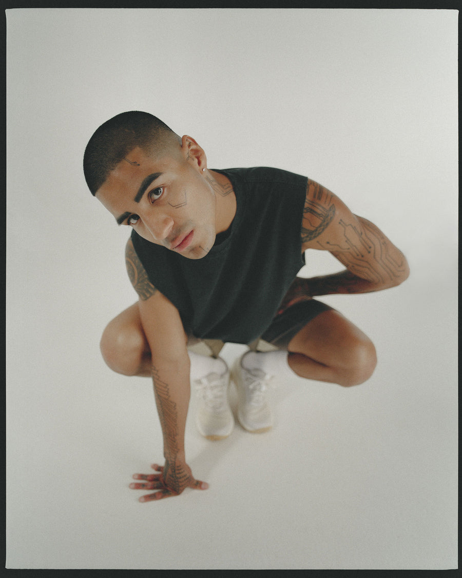 A man in a black sleeveless tee crouches and looks into the camera.