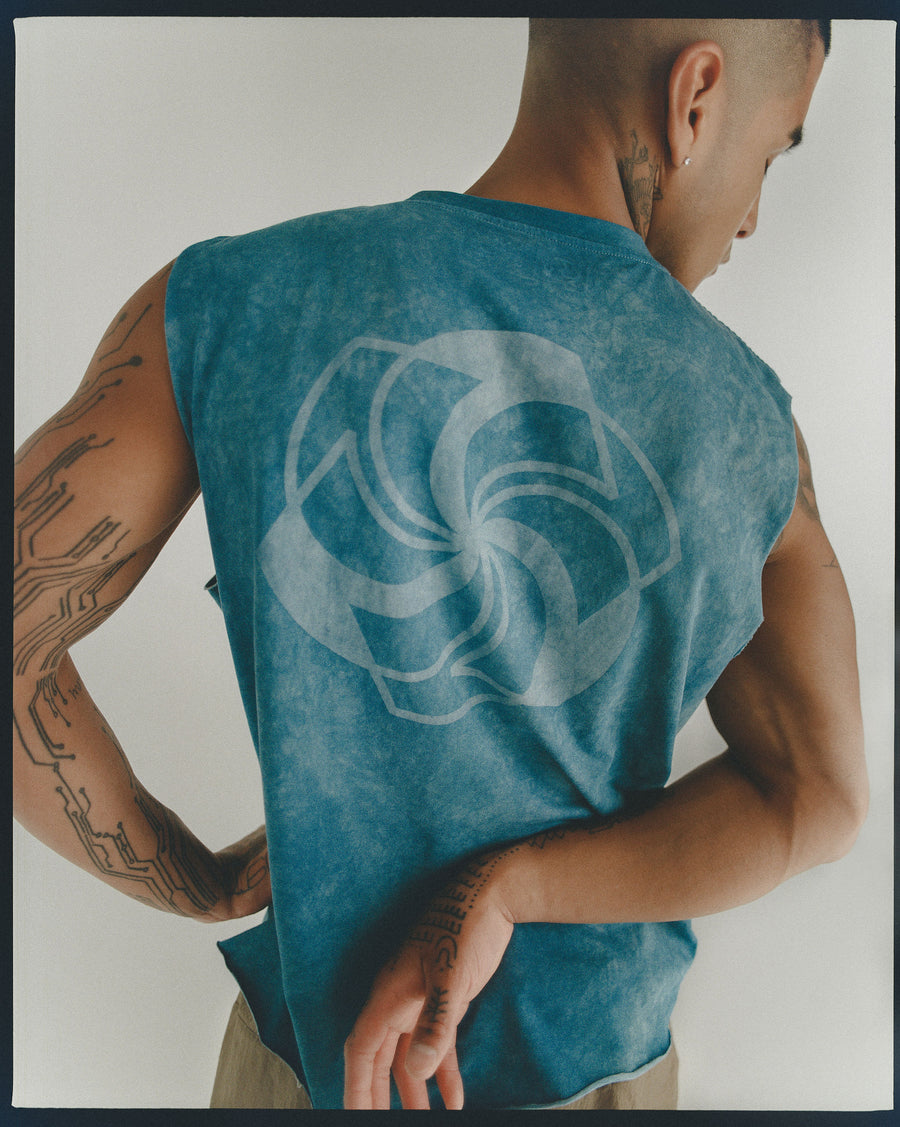 A man in a blue cutoff tee poses with his arm behind his back.