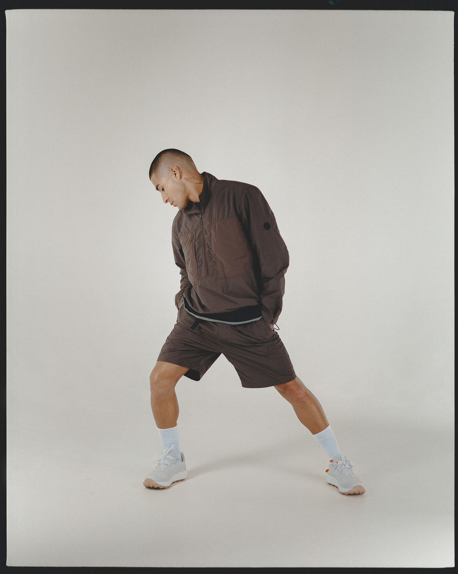 A man in a brown nylon pullover and brown shorts stretches.