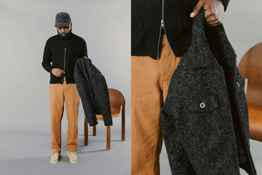 A man in a black sweater and sienna pants holds a black jacket in his hands.