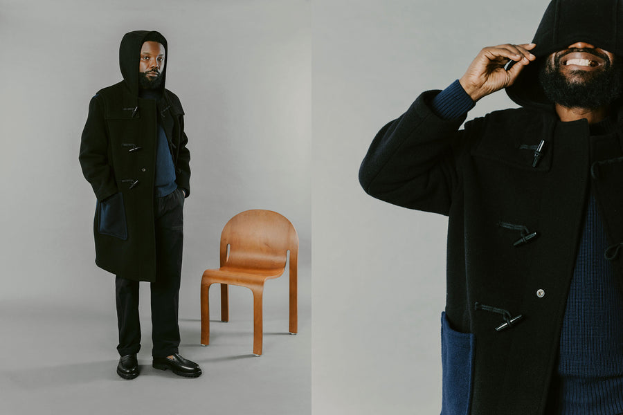 A man in a duffel coat stands in front of a chair.