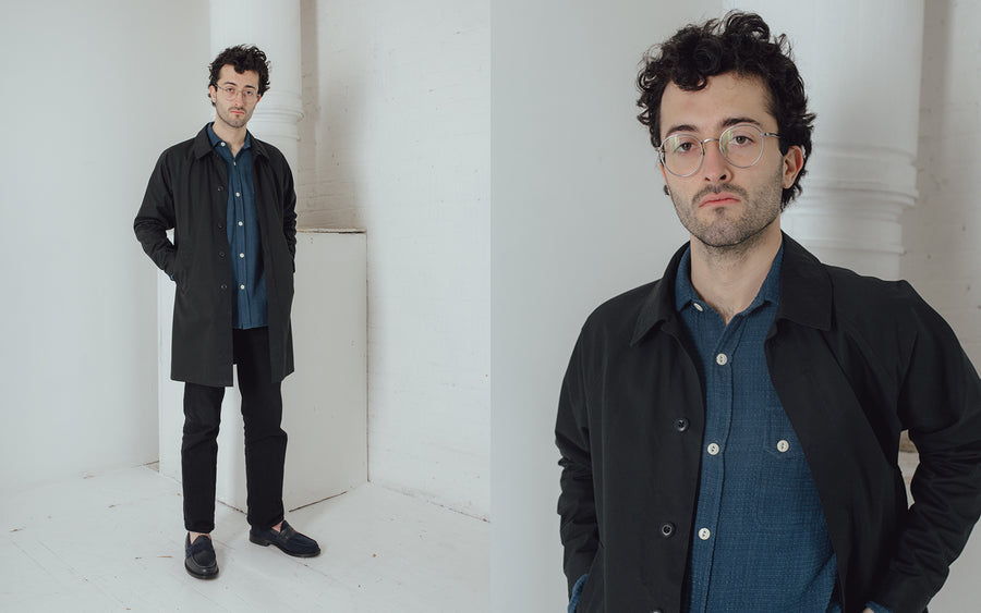 A man wears a black mac coat over an indigo grid shirt.