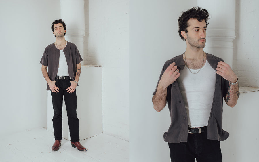 A man in a grey baseball shirt and black jeans looks into the camera.