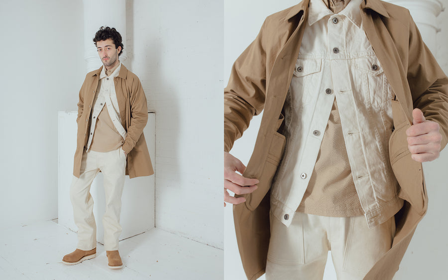 A man wears a tan mac coat over a natural denim jacket and fatigue pants.