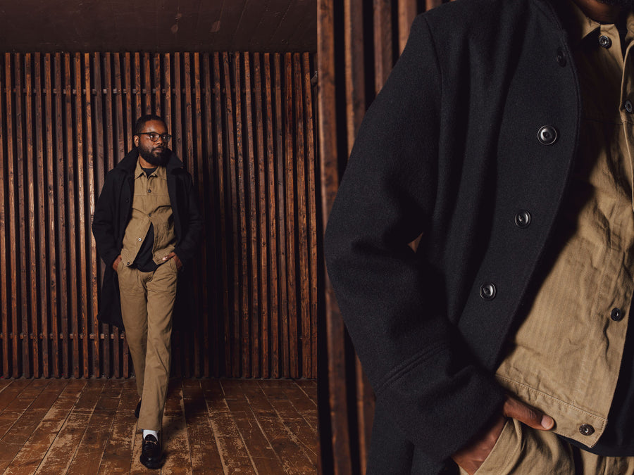 A man in a long black overcoat and tan pants stands against a wooden slat wall.