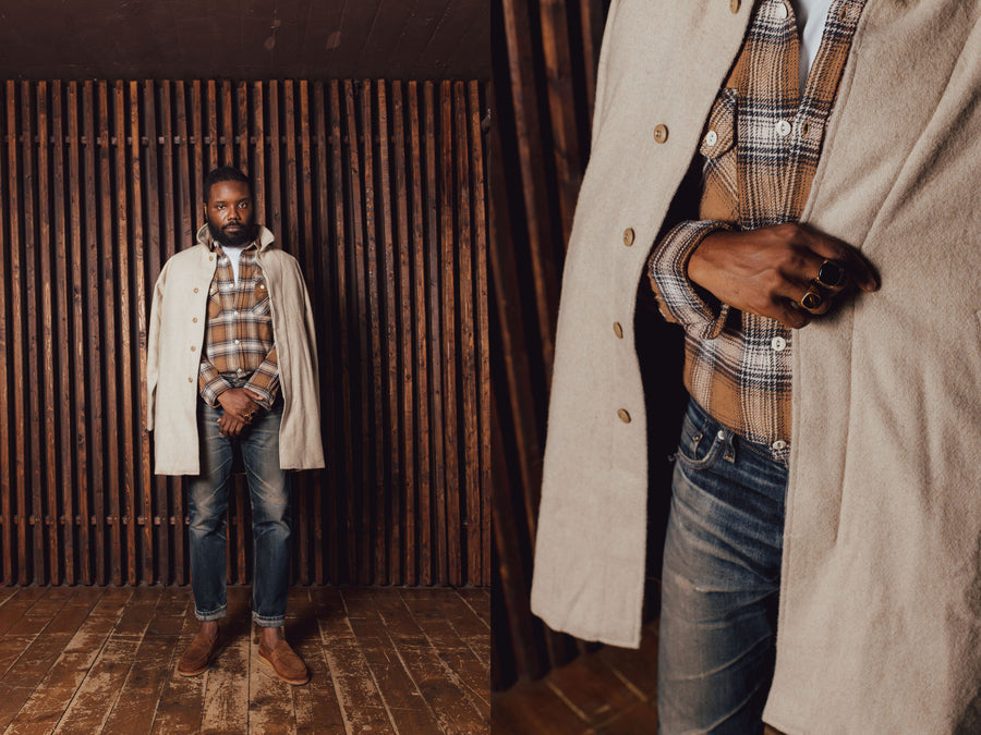 A man in a light tan wool coat stands in front of a wooden slat wall.