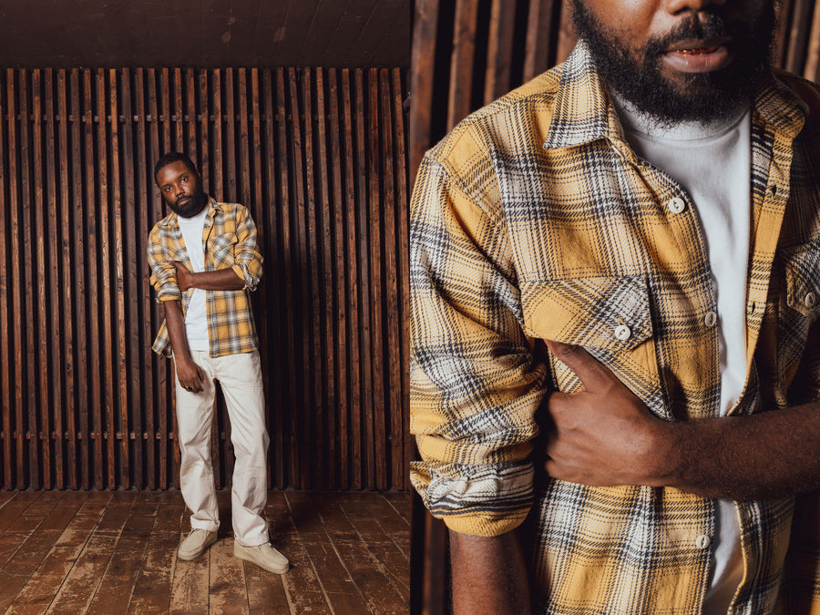 A man in a yellow flannel and white pants stands in front of a wood slat wall.