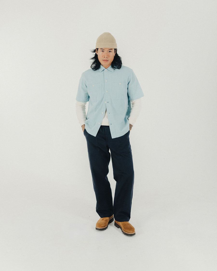 A man in a tan knit cap and light blue shirt poses for the camera with his hands in his pockets.