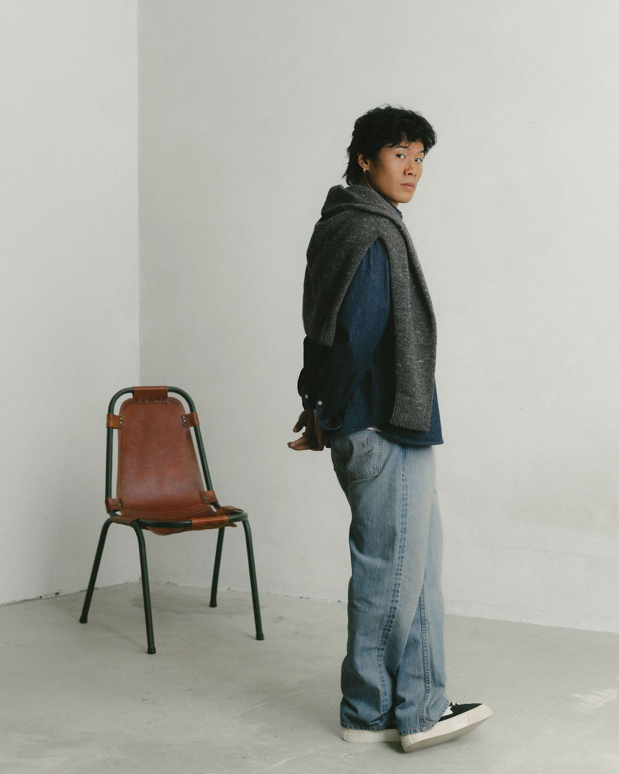 A man in a denim shirt with a sweater over his shoulders poses.