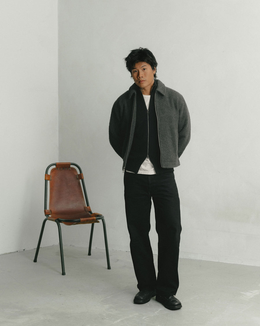 A man in a grey wool cropped jacket layered over a black sweater poses and looks into the camera.