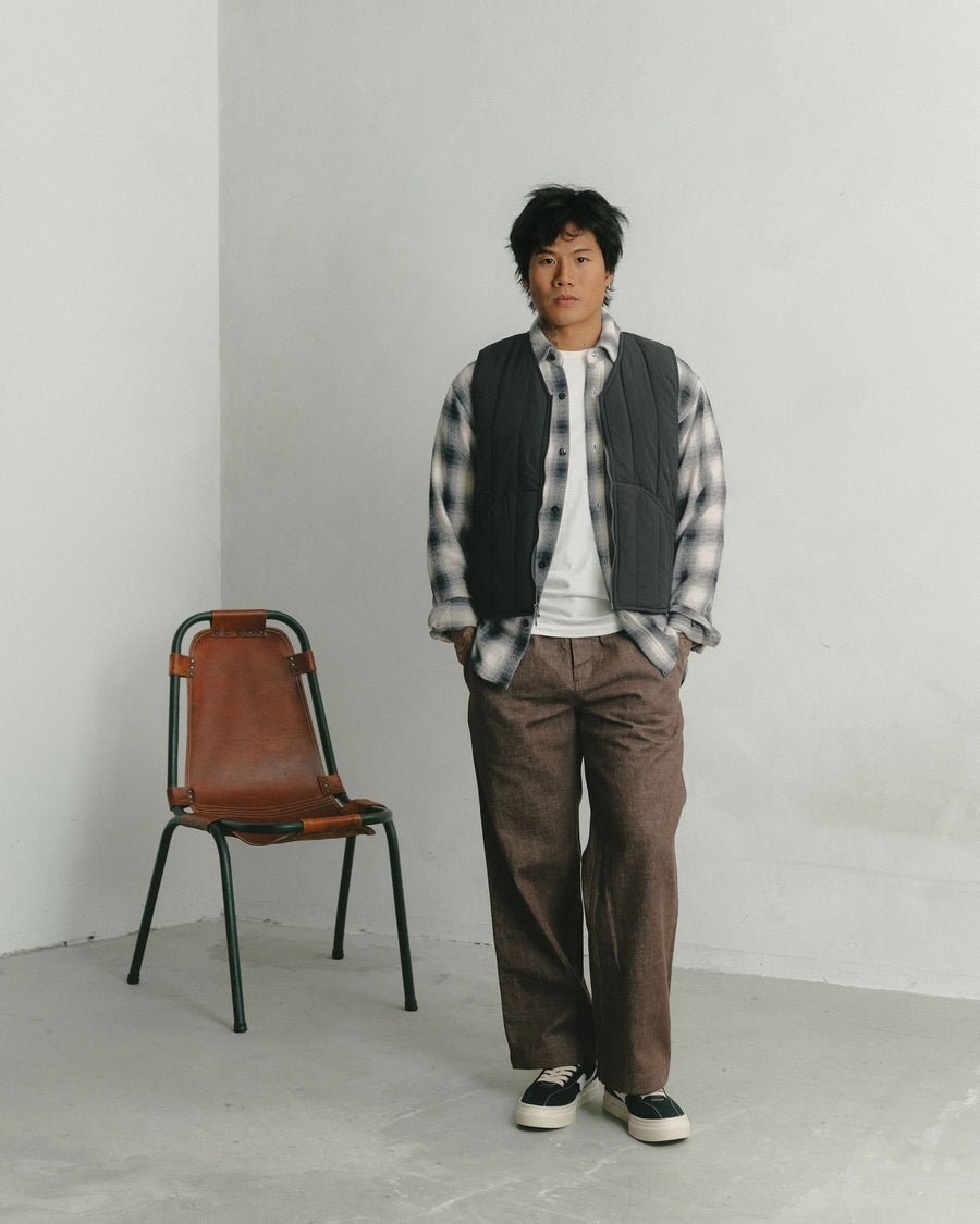 A man in a black and white flannel layered under a quilted black vest poses with his hands in his pockets.