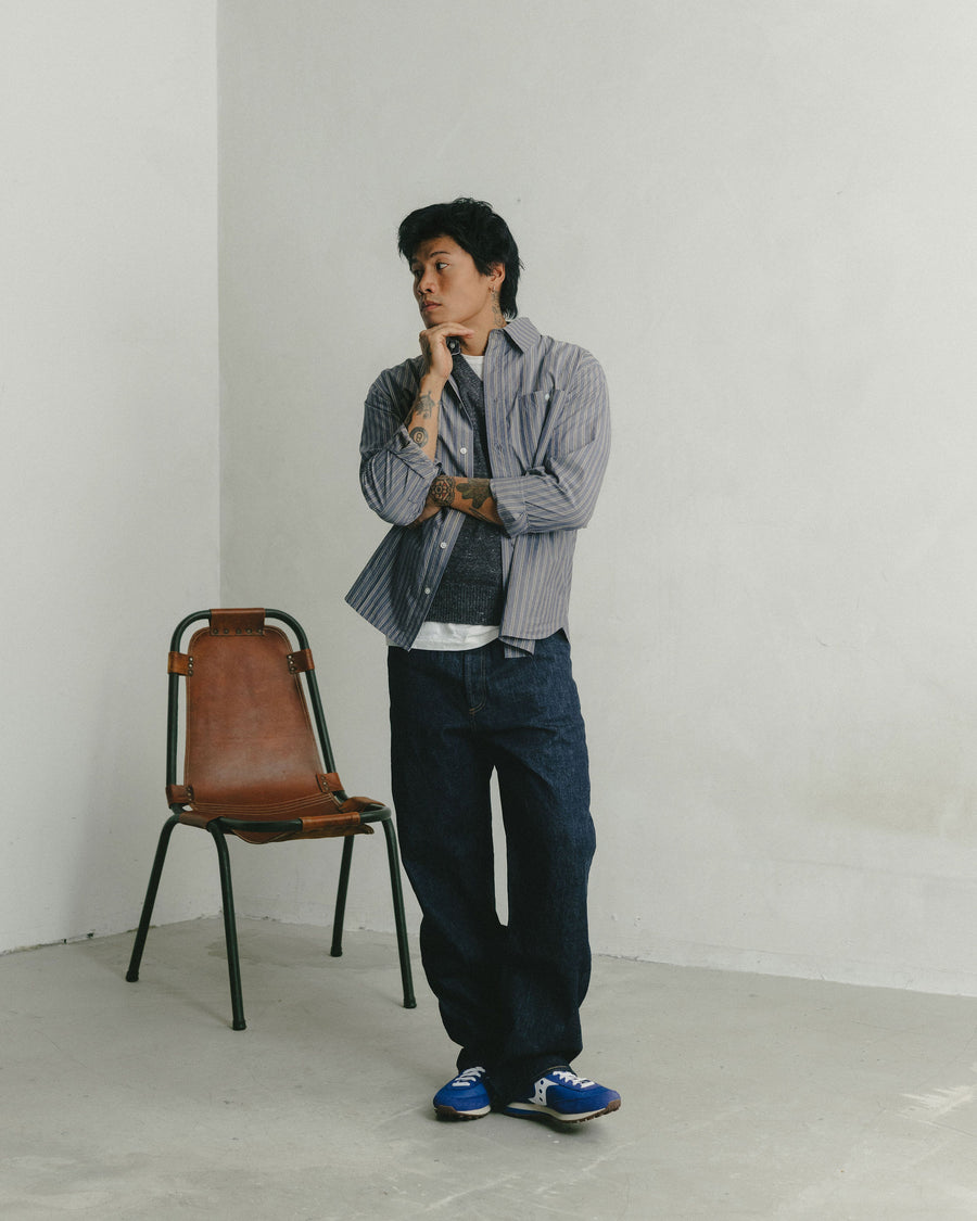 A man in a striped blue poplin shirt layered over a grey sweater vest poses and looks off to the side.