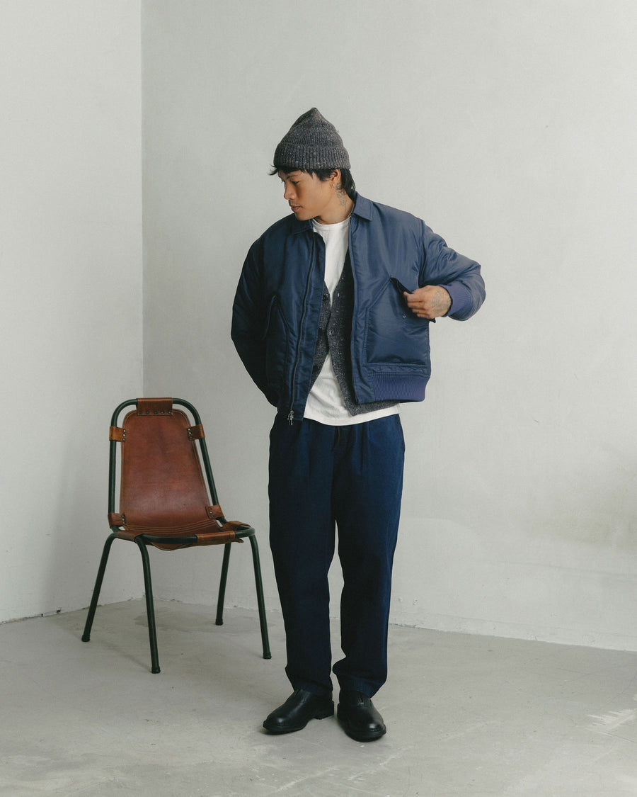 A man in a satin blue bomber jacket and dark indigo pants reaches into his pocket.