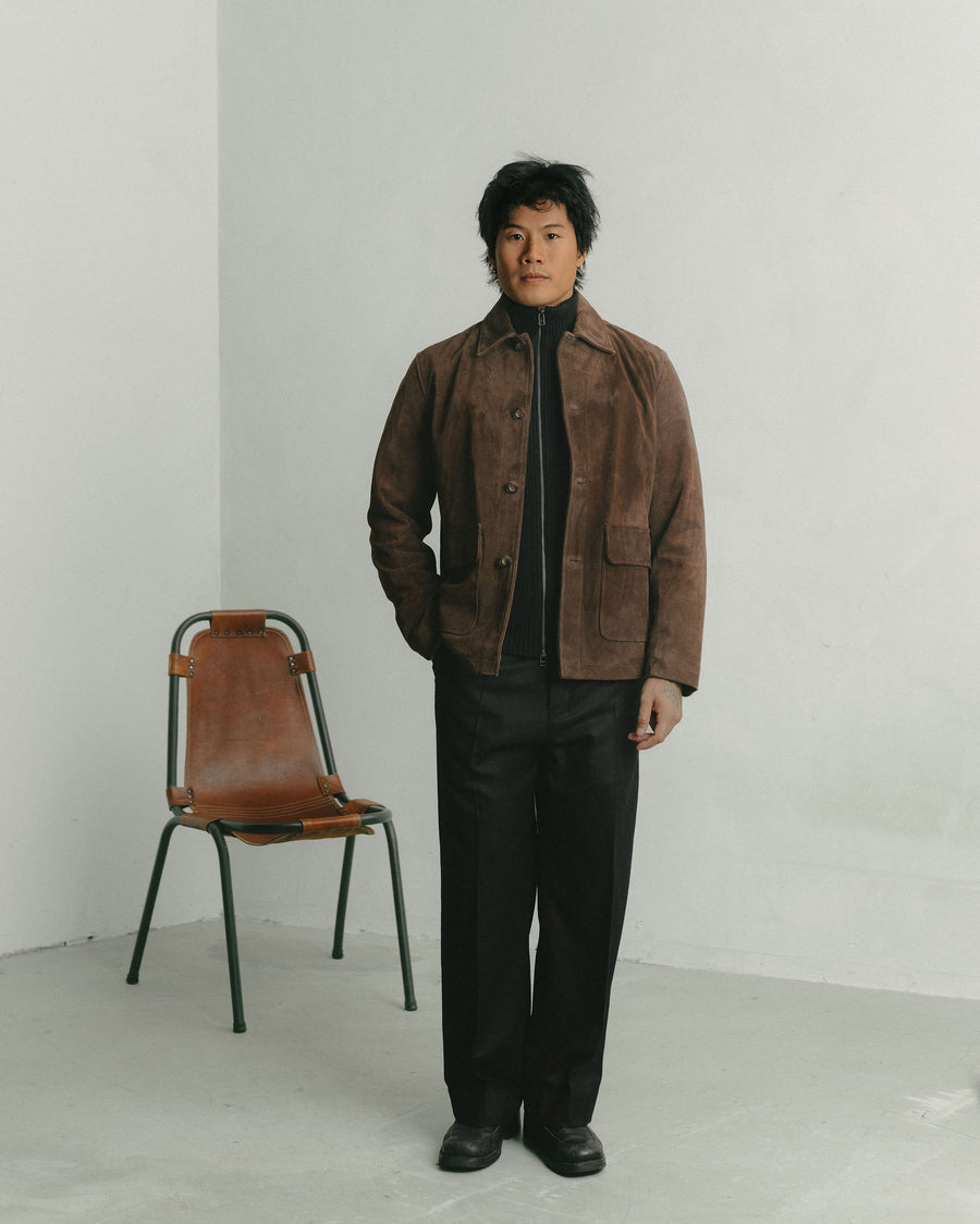 A man in a brown suede jacket and black wool pants stands in front of a leather chair.
