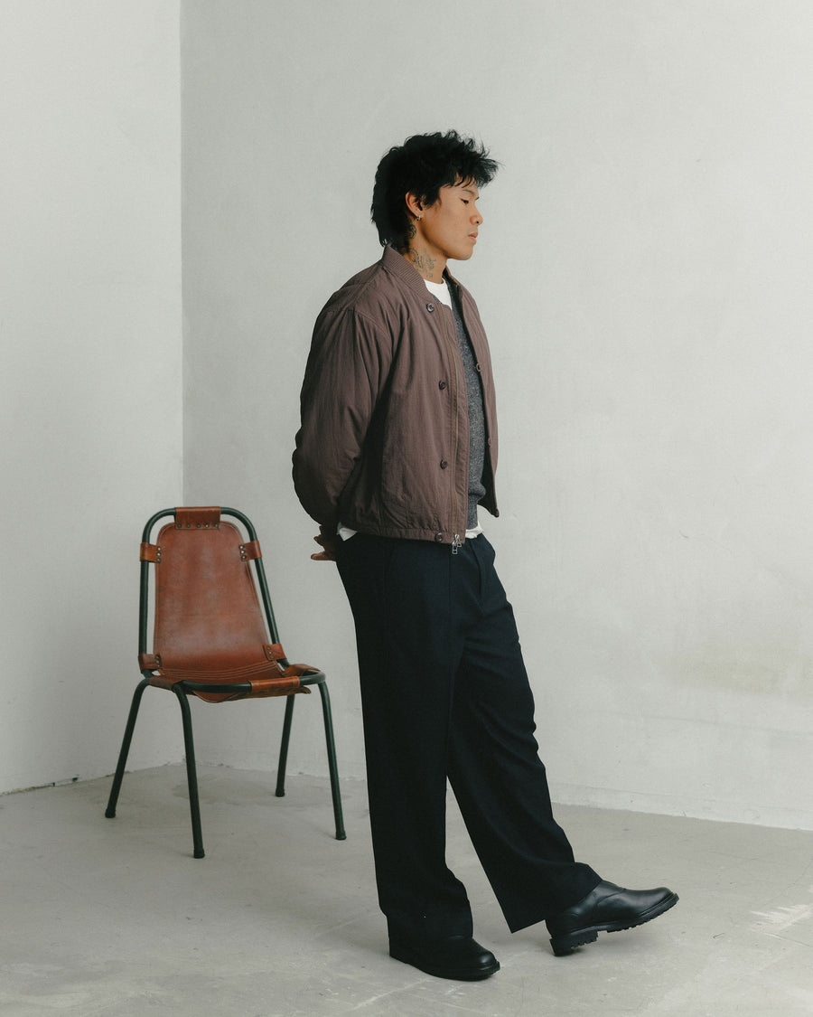 A man in a brown bomber jacket and black wool pants paces against a concrete backdrop.