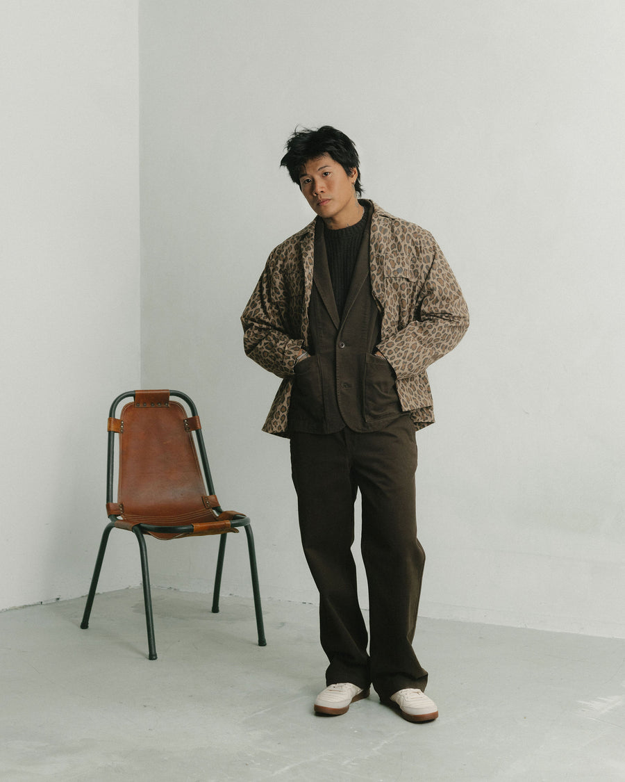 A man in a leopard print jacket layered over a chocolate brown cotton suit stands in front of a leather chair.