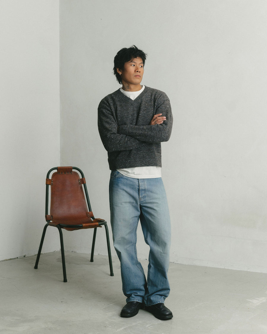 A man in a grey v-neck sweater and light wash jeans with arms akimbo.