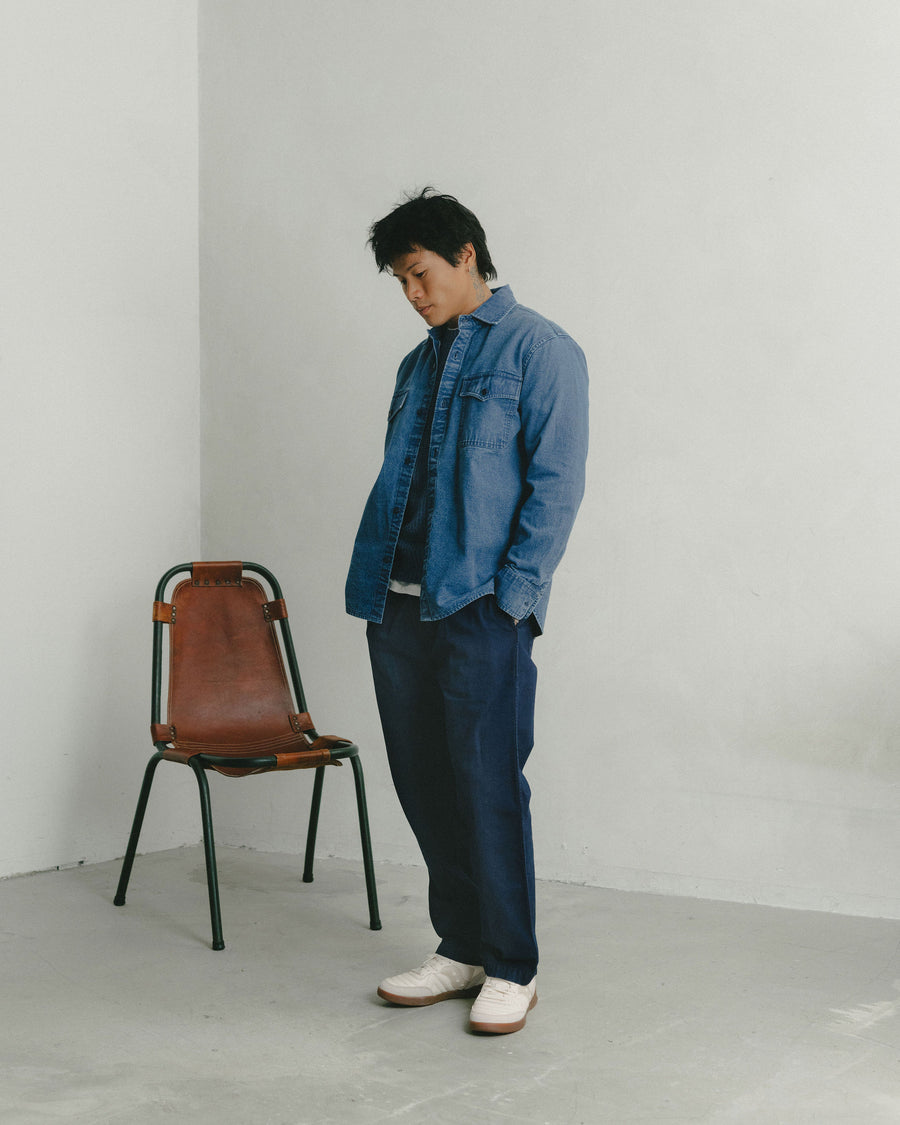 A man in a washed-out blue canvas shirt worn over dark indigo pants looks down at the floor.
