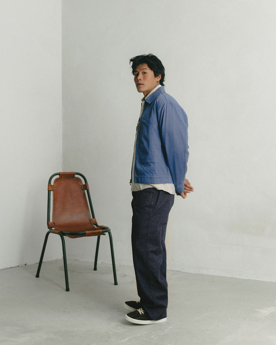 A man in a light blue cropped jacket worn over dark indigo pants poses with his side to the camera.