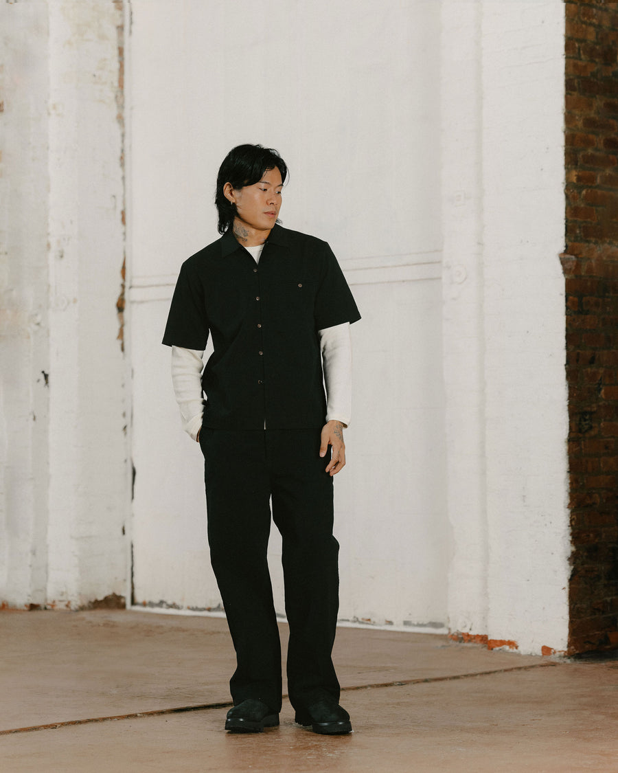 A man in a black short sleeve button down layered over a white thermal poses.