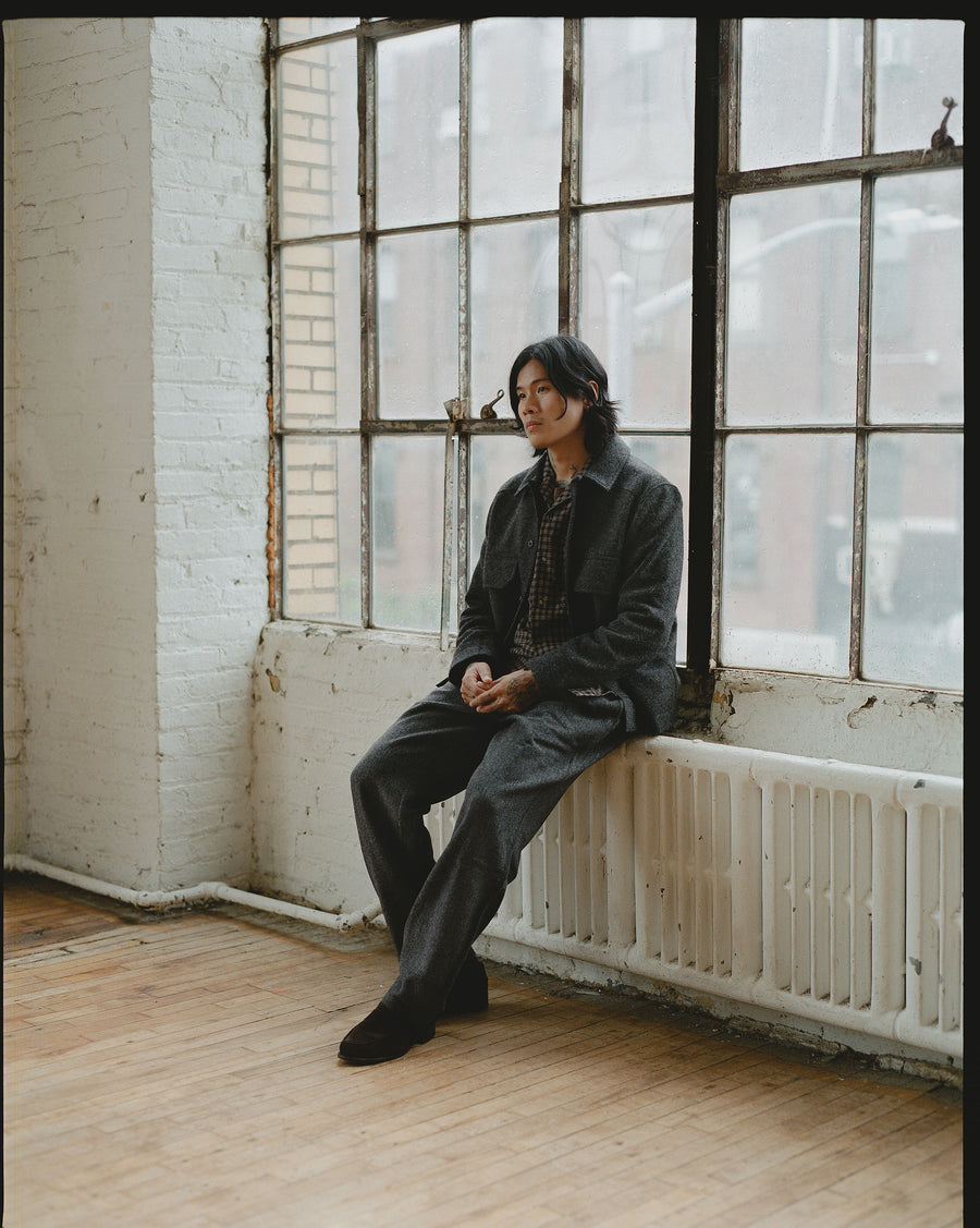 A man in a grey wool shirt and wool pants sits on a windowsill.