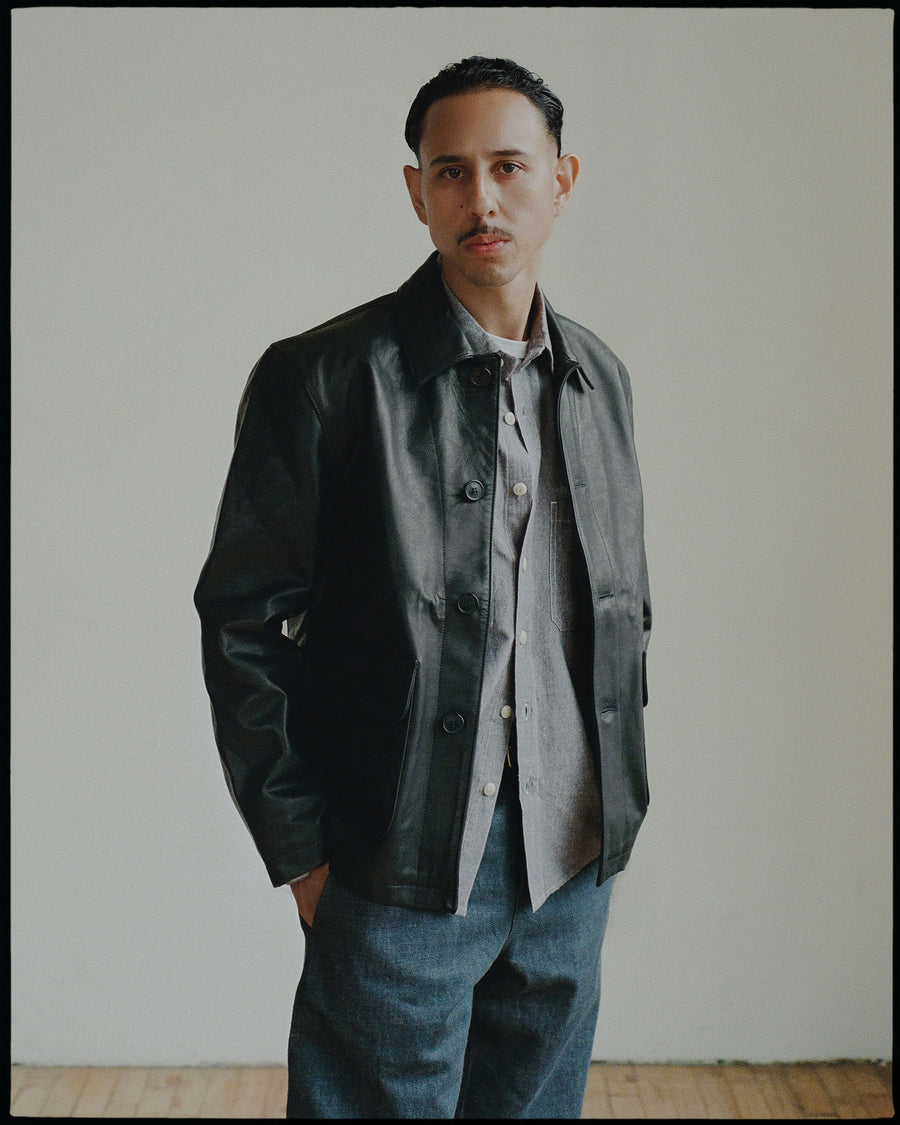 A man in a leather jacket and grey chambray looks into the camera.