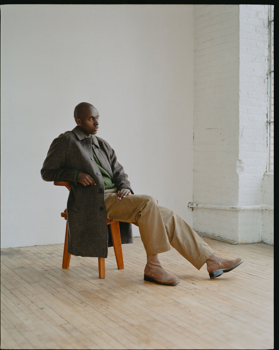 A man in a long wool overcoat and green sweater sits in a wooden chair.