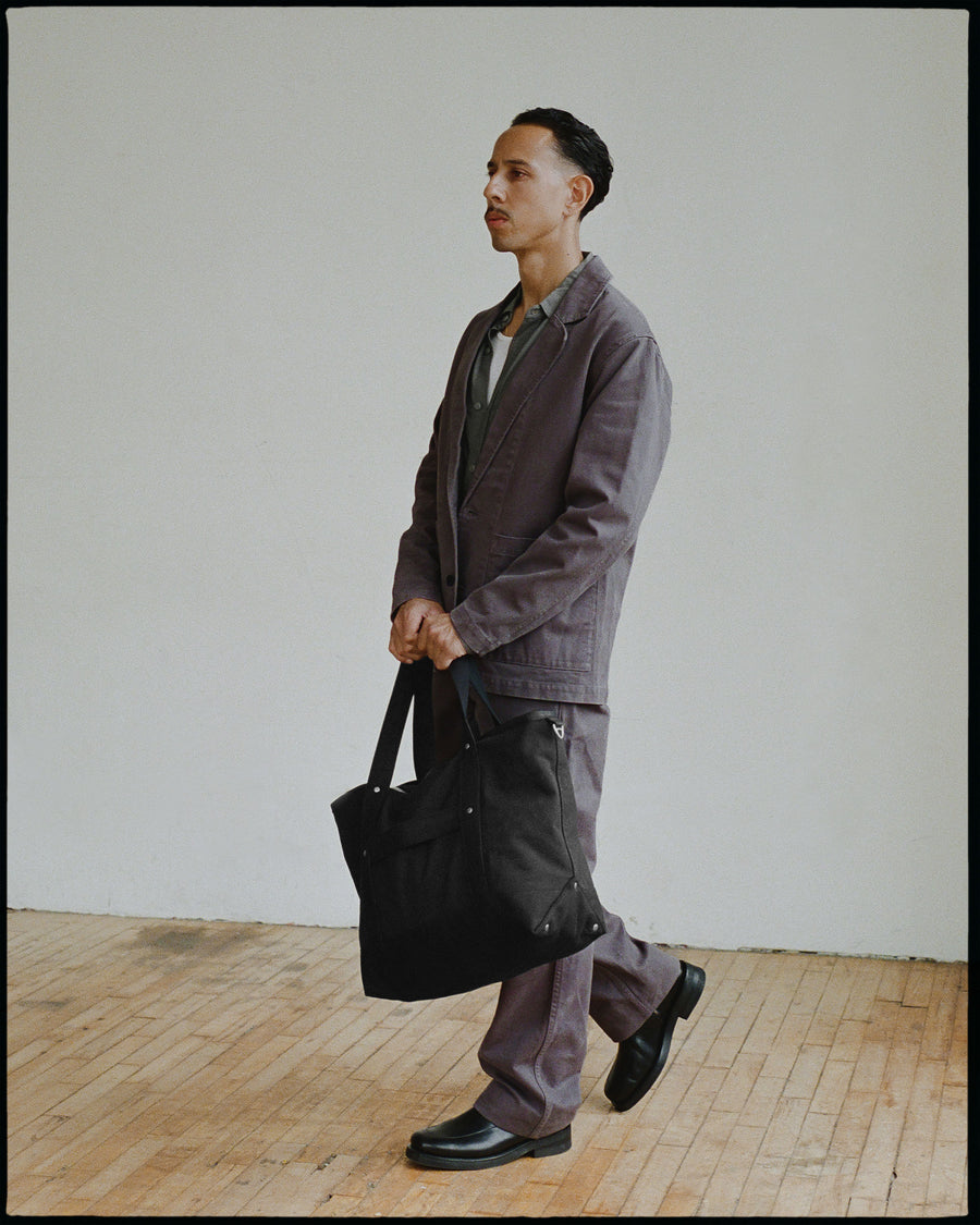 A man in a charcoal grey suit holding a black tote bag.