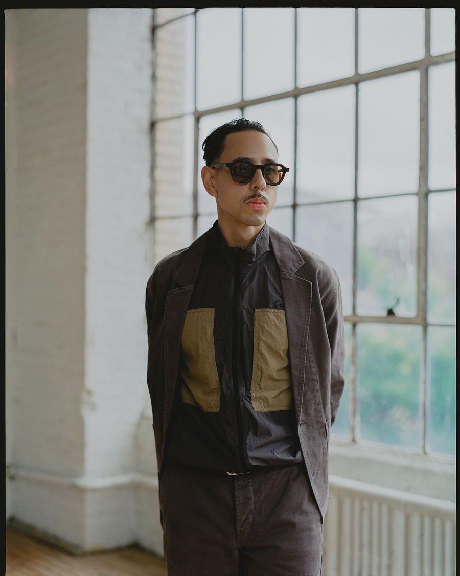 A man in a track jacket with a blazer over it stands in front of a window.