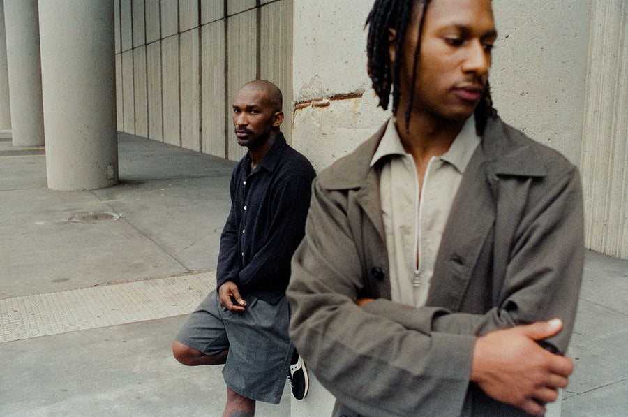 Two men wearing olive and black clothes stand in front of a concrete pillar.