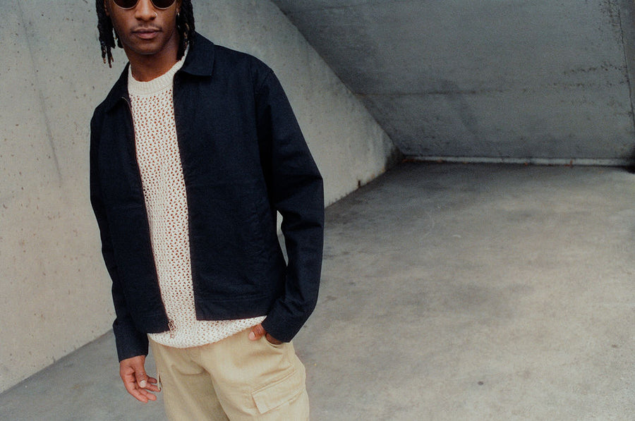 A man in a white knit tee and cropped black jacket stands on concrete.