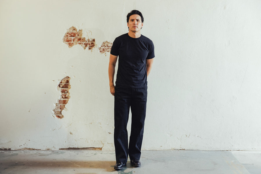 A man in a black tee and black jeans stands in front of a wall.