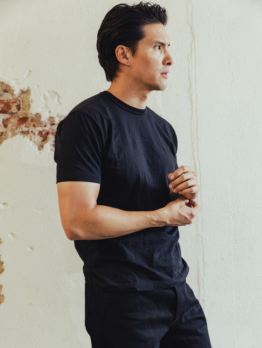 A man in a black tee and black jeans stands by a white wall.