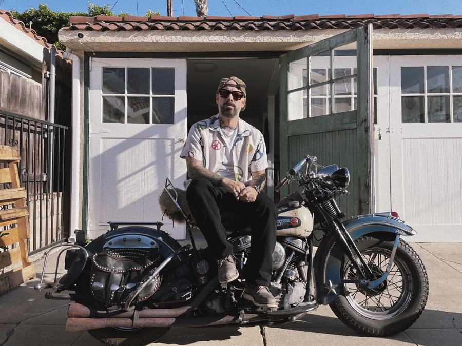 A man in a multicolor peace shirt on a vintage motorcycle.