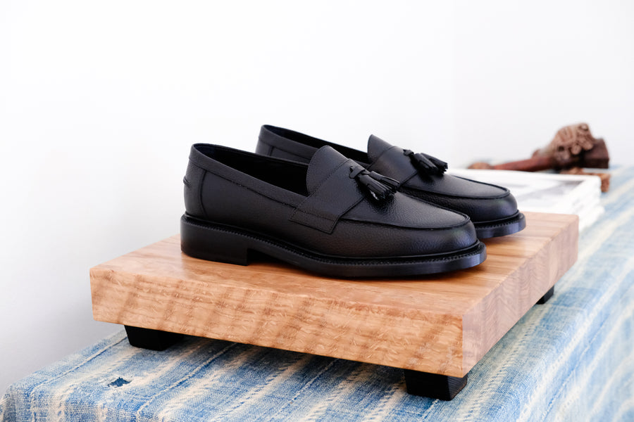 A pair of black pebbled tassel loafers on a wooden block stand.