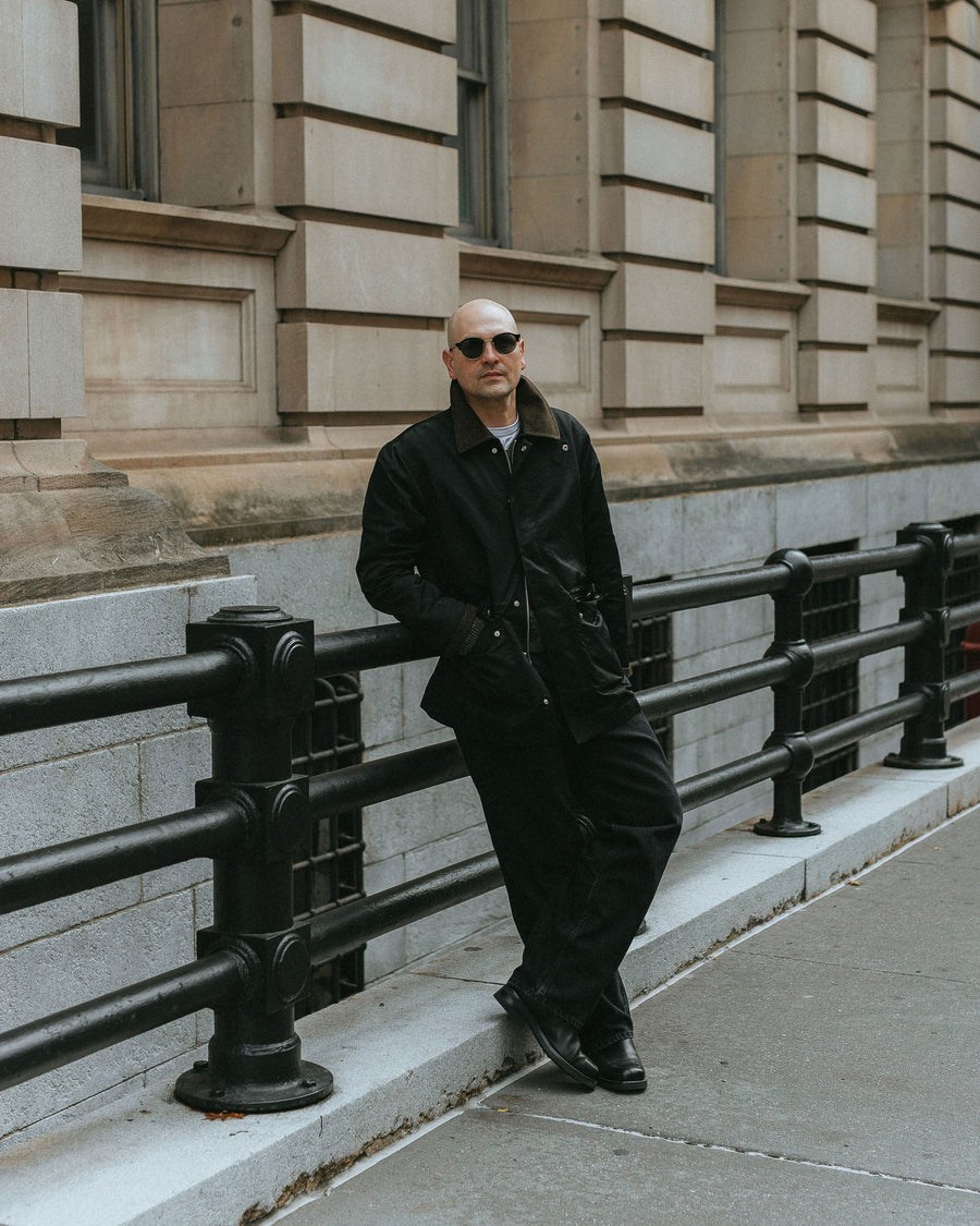 A man in black sunglasses and black nylon coat leans against some railing.