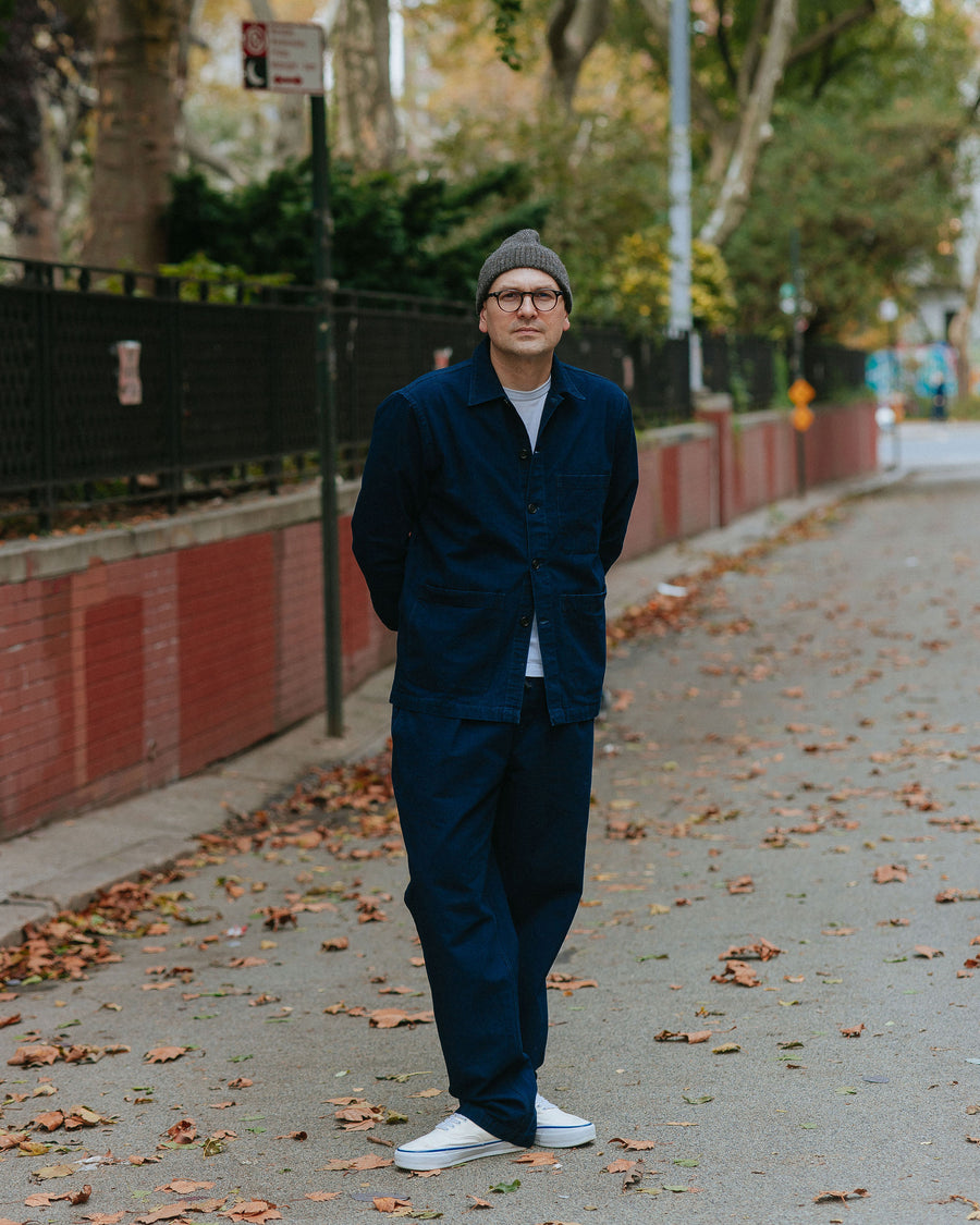 A man in a navy blue two piece set stands amongst leaves.