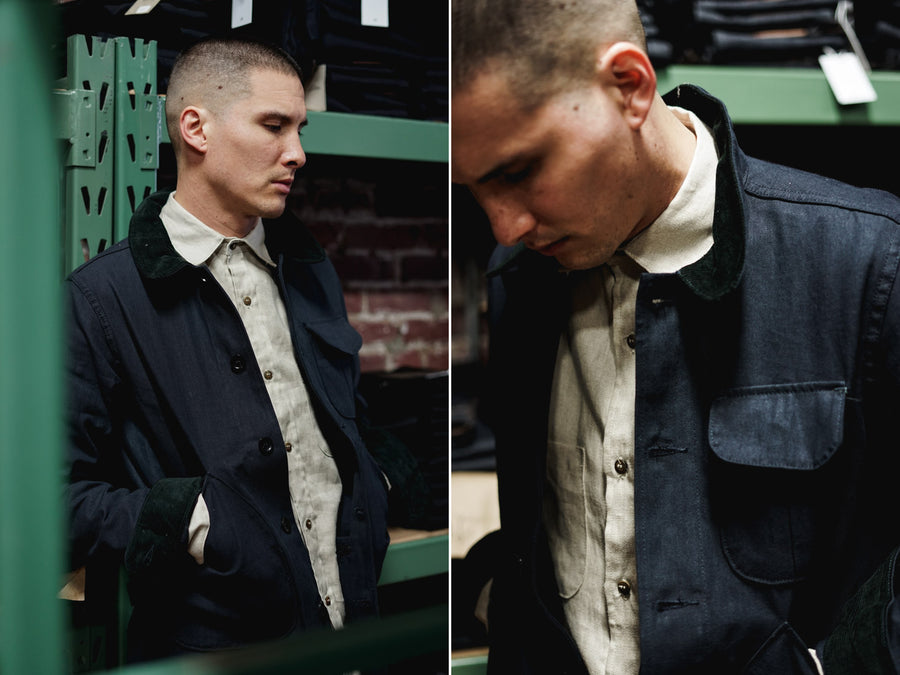 The model wears a 3sixteen Hunting Jacket in navy and a 3sixteen Linen Shirt in natural, while leaning against a rack.