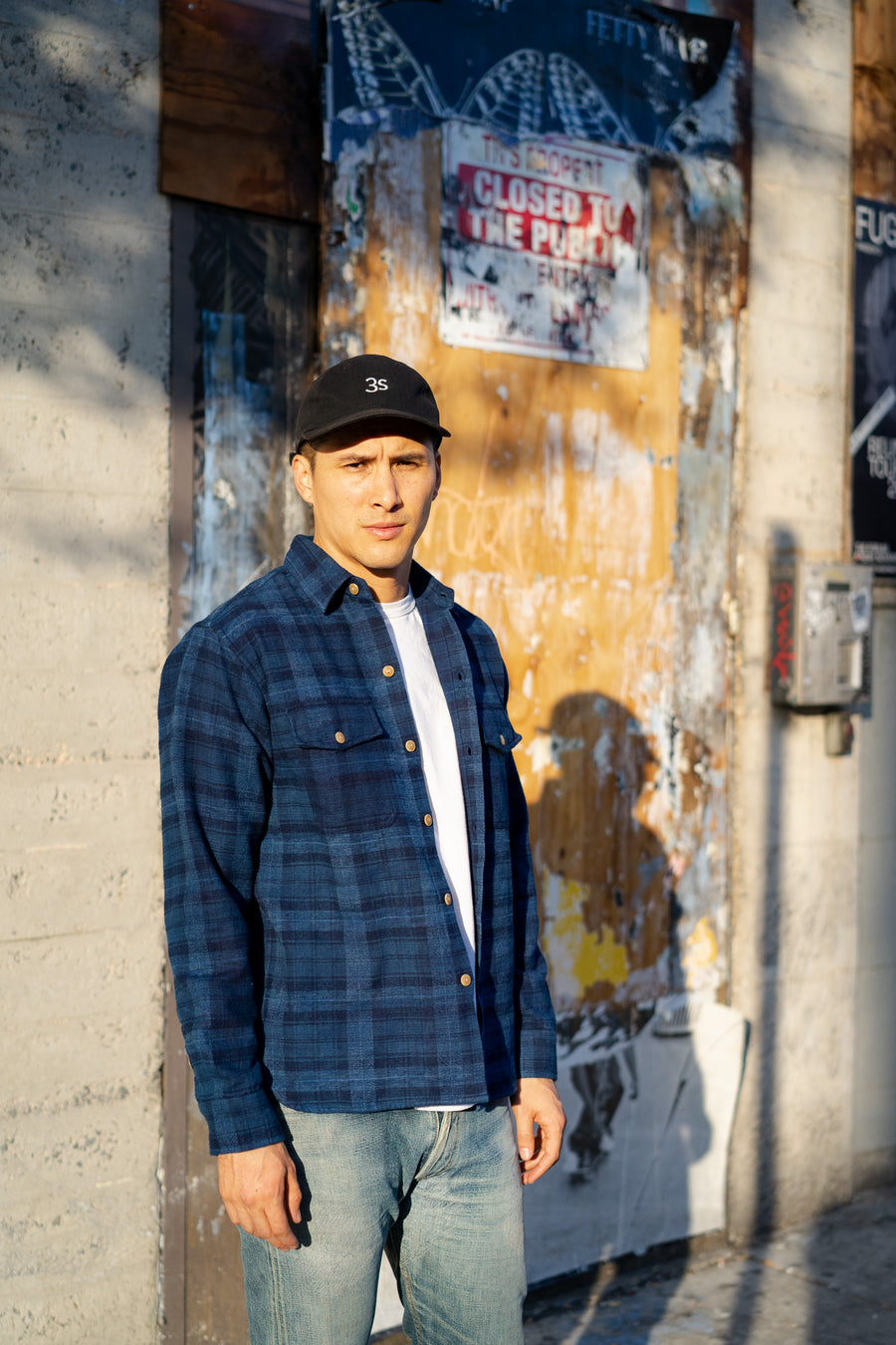 A man stands on a street wearing a blue flannel.