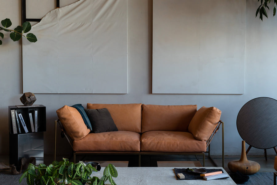 A beautiful tan couch sits in a large room with plants and other home accessories surrouunding it.