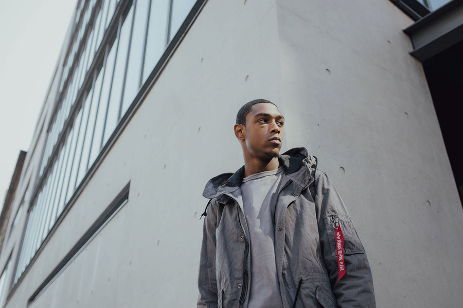 A young man wears the Alpha Industries grey fishtail parka in front of a concrete building