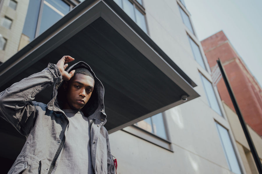 A young man wears the Alpha Industries grey fishtail parka in front of a concrete building