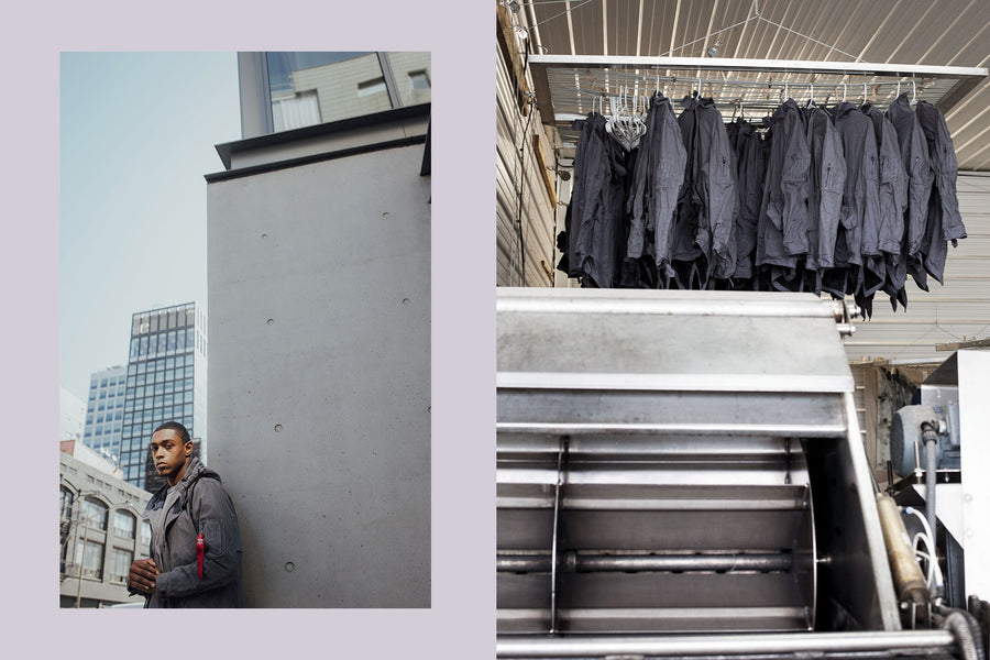 A man stands by a concrete building wearing a grey Alpha Industries jacket.