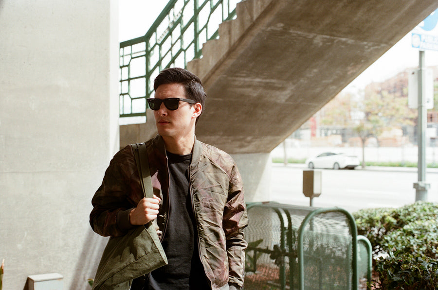 A man in an olive bomber and tote looks left.