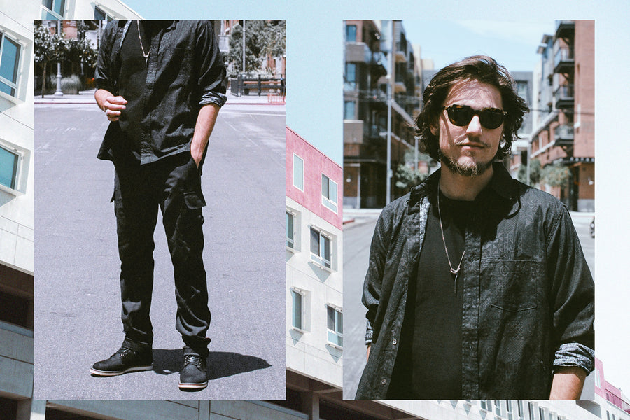 A man models sunglasses and a black outfit and black boots.