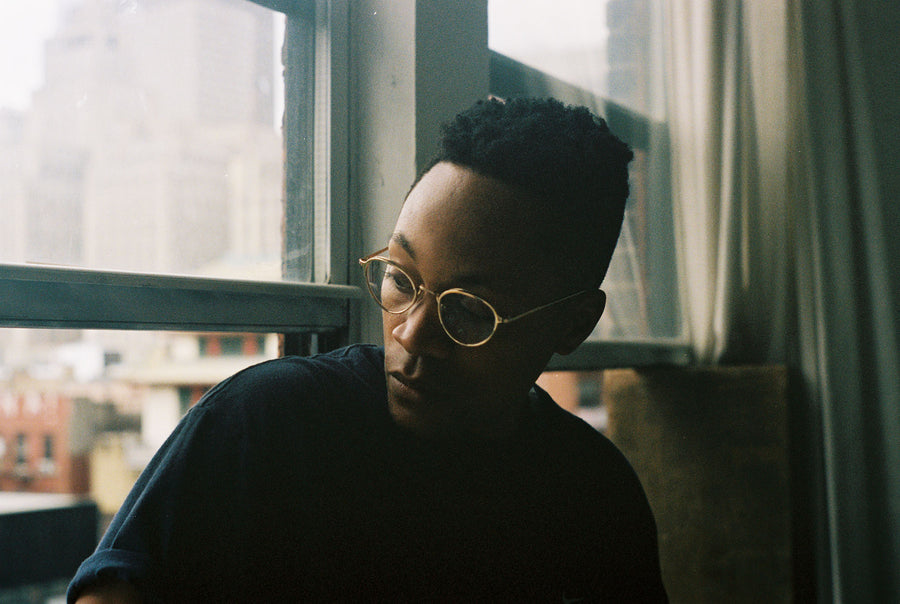 A man with gold-rimmed glasses sits perched on a windowsill