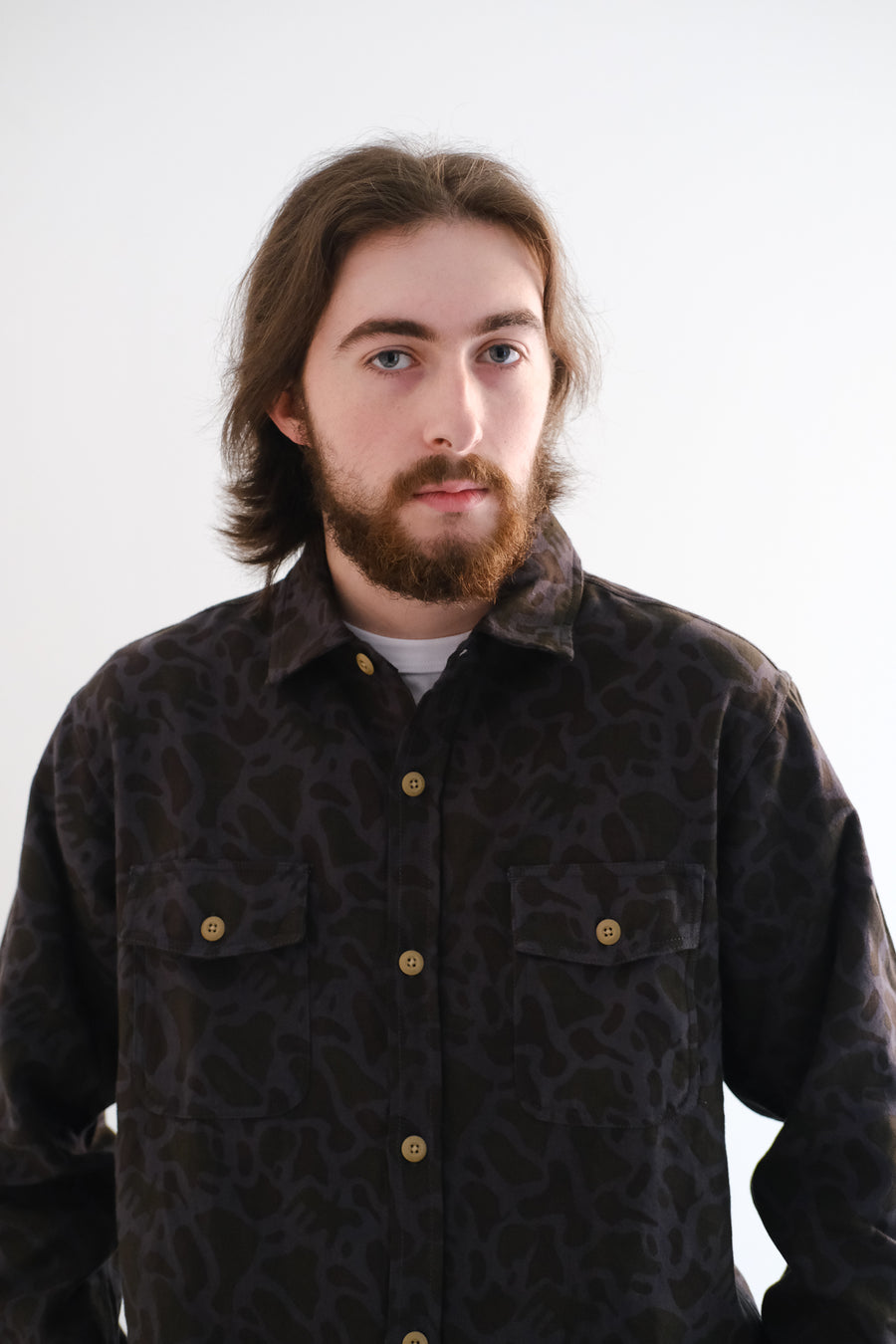 A man with long brown hair wears a black camouflage shirt.